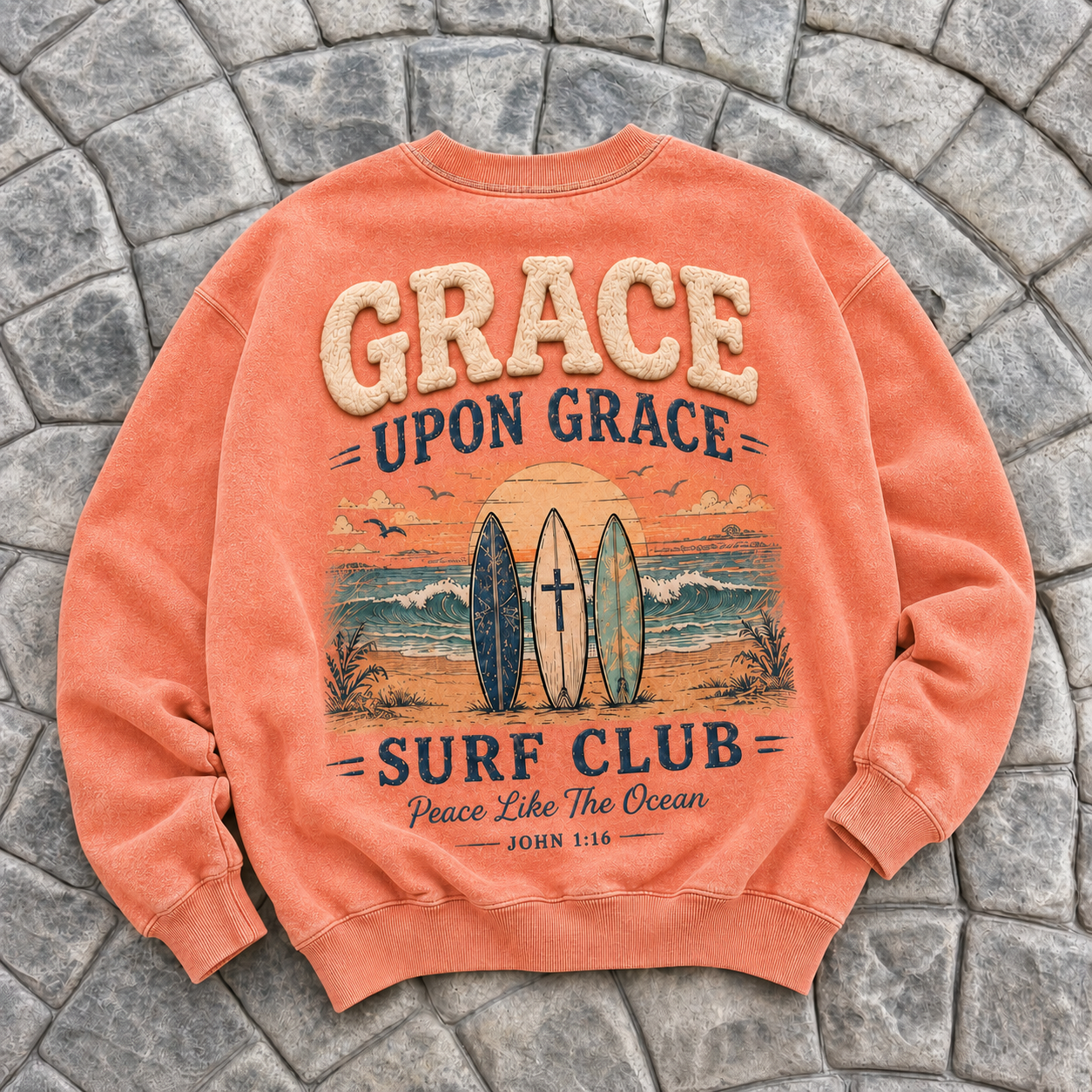 Coral-colored crewneck with 'Grace Upon Grace Surf Club' design on a stone surface