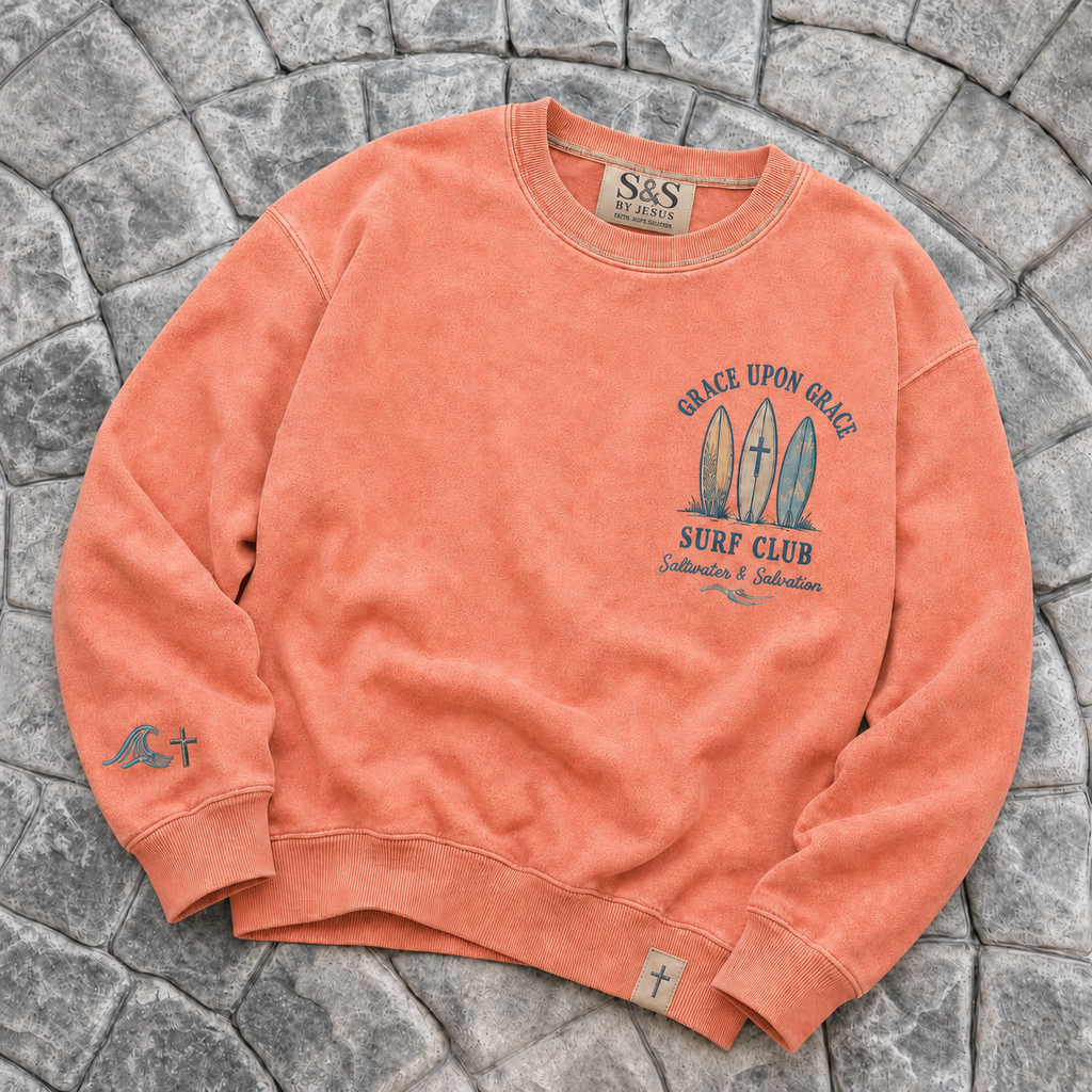 Coral-colored crewneck with 'Grace Upon Grace Surf Club' design on a stone surface