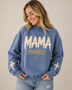 Woman wearing a blue sweatshirt with 'MAMA PRAYED' text and crosses on a beige background