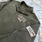 Detail view of heritage olive Saved by Jesus quarter zip with Strength Found in Christ sleeve patch and dark gunmetal cross zipper pull
