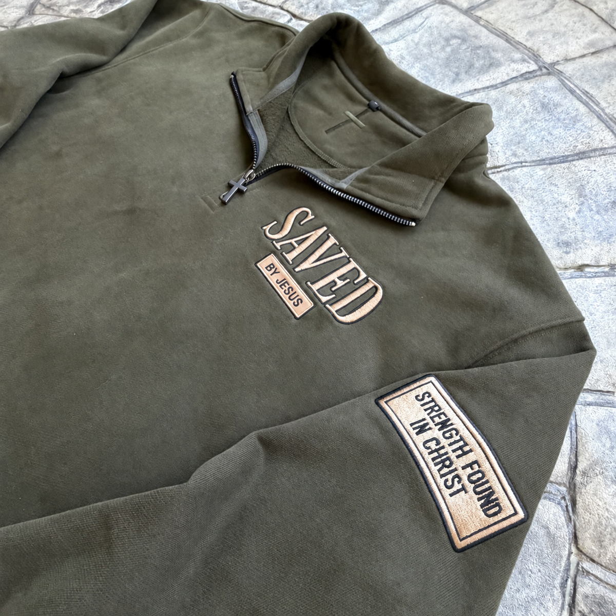 Detail view of heritage olive Saved by Jesus quarter zip with Strength Found in Christ sleeve patch and dark gunmetal cross zipper pull